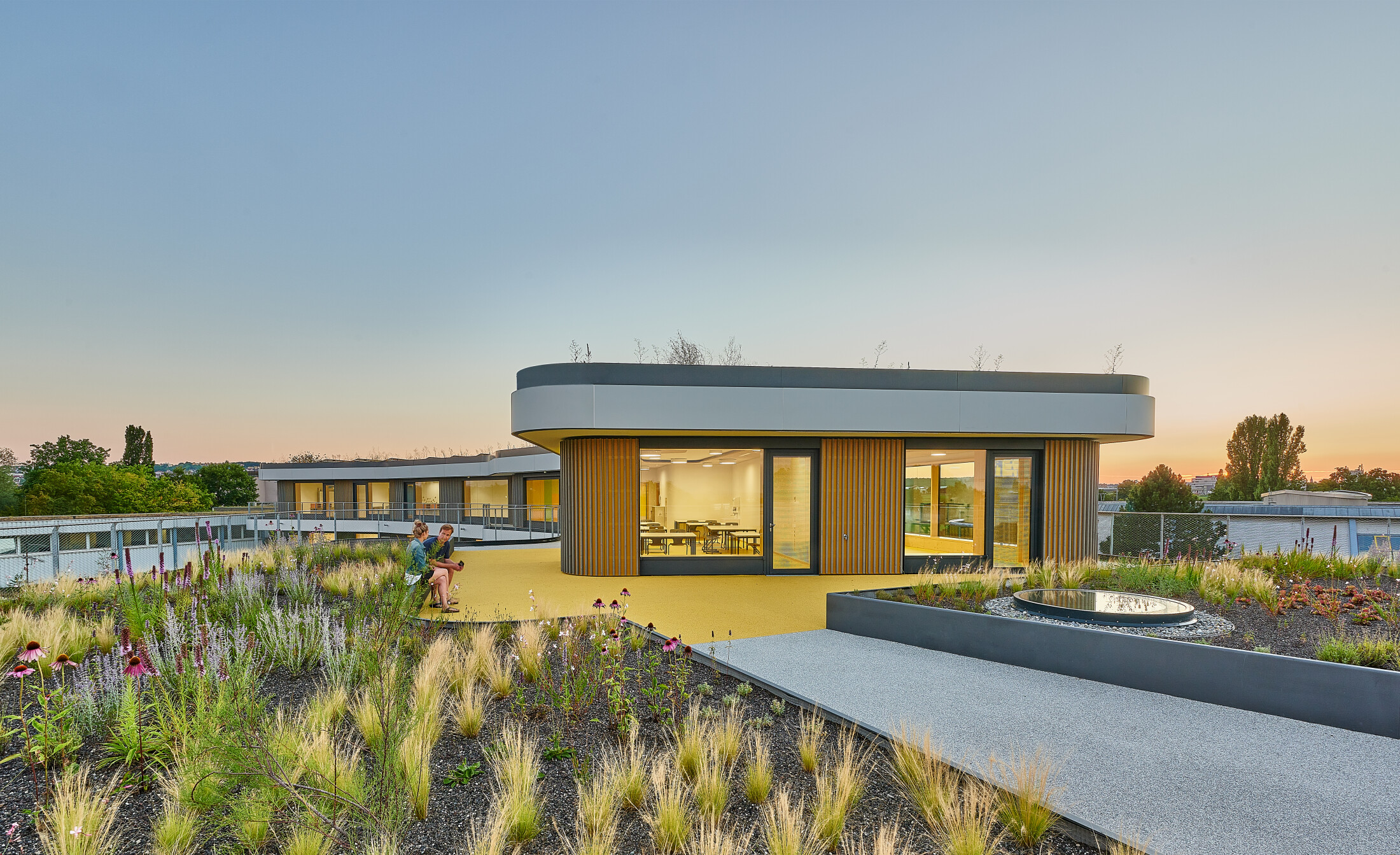 Project by Behnisch Architekturbuero, Gotthard-Müller-School Bernhausen. A rooftop terrace with a landscaped garden and pathways; two people sit next to a modern, circular building with large windows at sunset. Part of the flooring is marked in yellow.