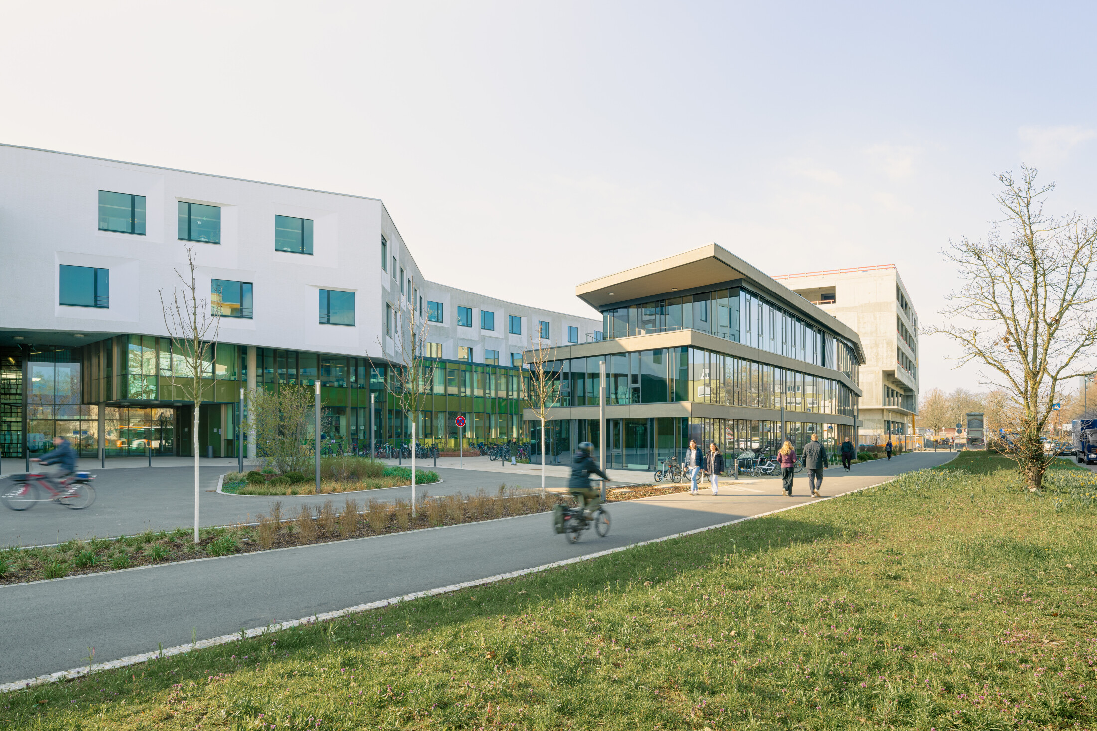 Project by Behnisch Architekturbuero, NCT New Building and Expansion. Modern campus buildings with glass facades, people walking and cycling on a path, and a grassy area with trees in the foreground.