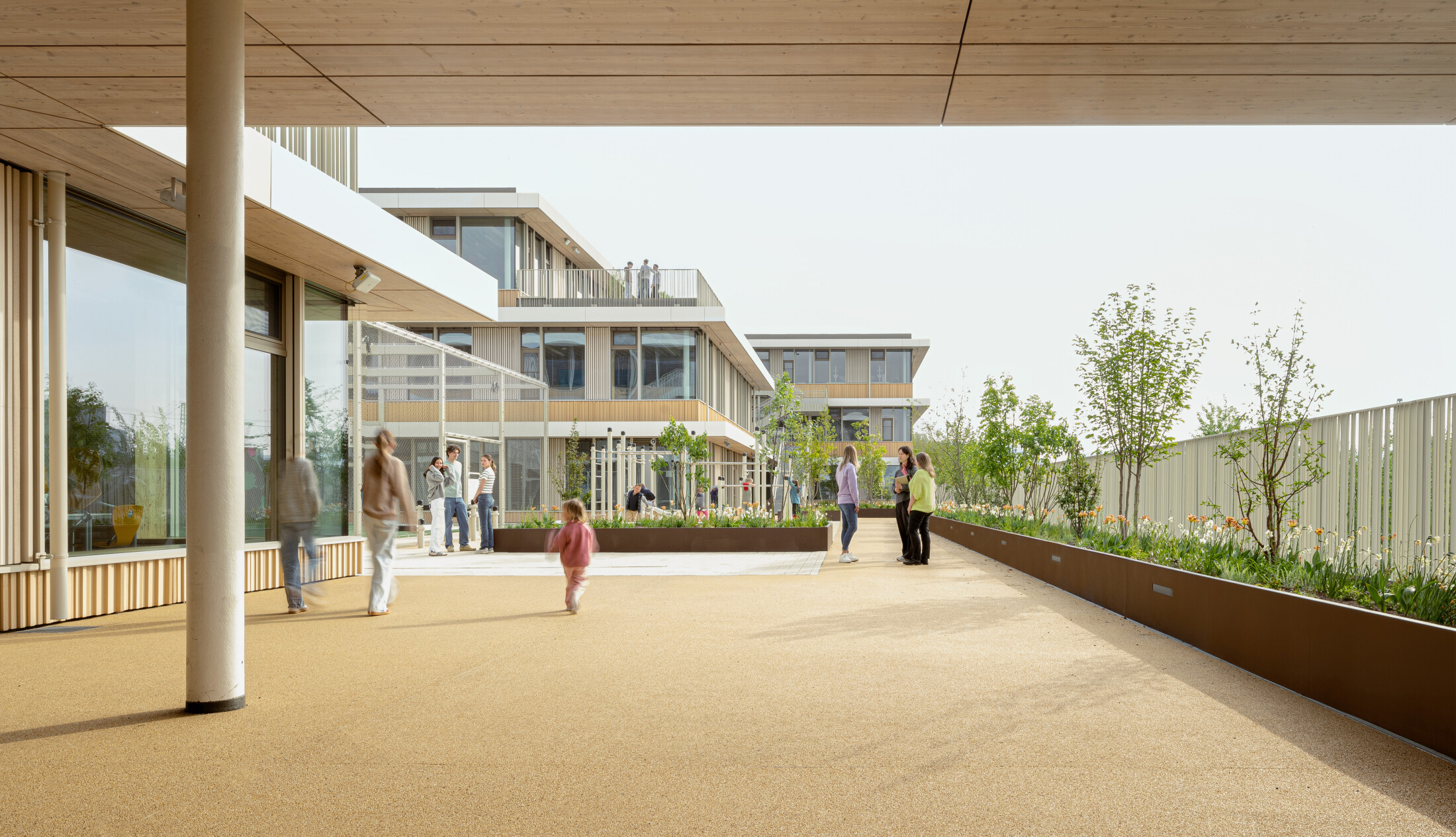 Project by Behnisch Architekturbuero, Josef-Schwarz School. Students and teachers are in a green outdoor area between the modern buildings. A raised bed with plants and trees can be seen on the right. A climbing frame is visible in the background on the left.