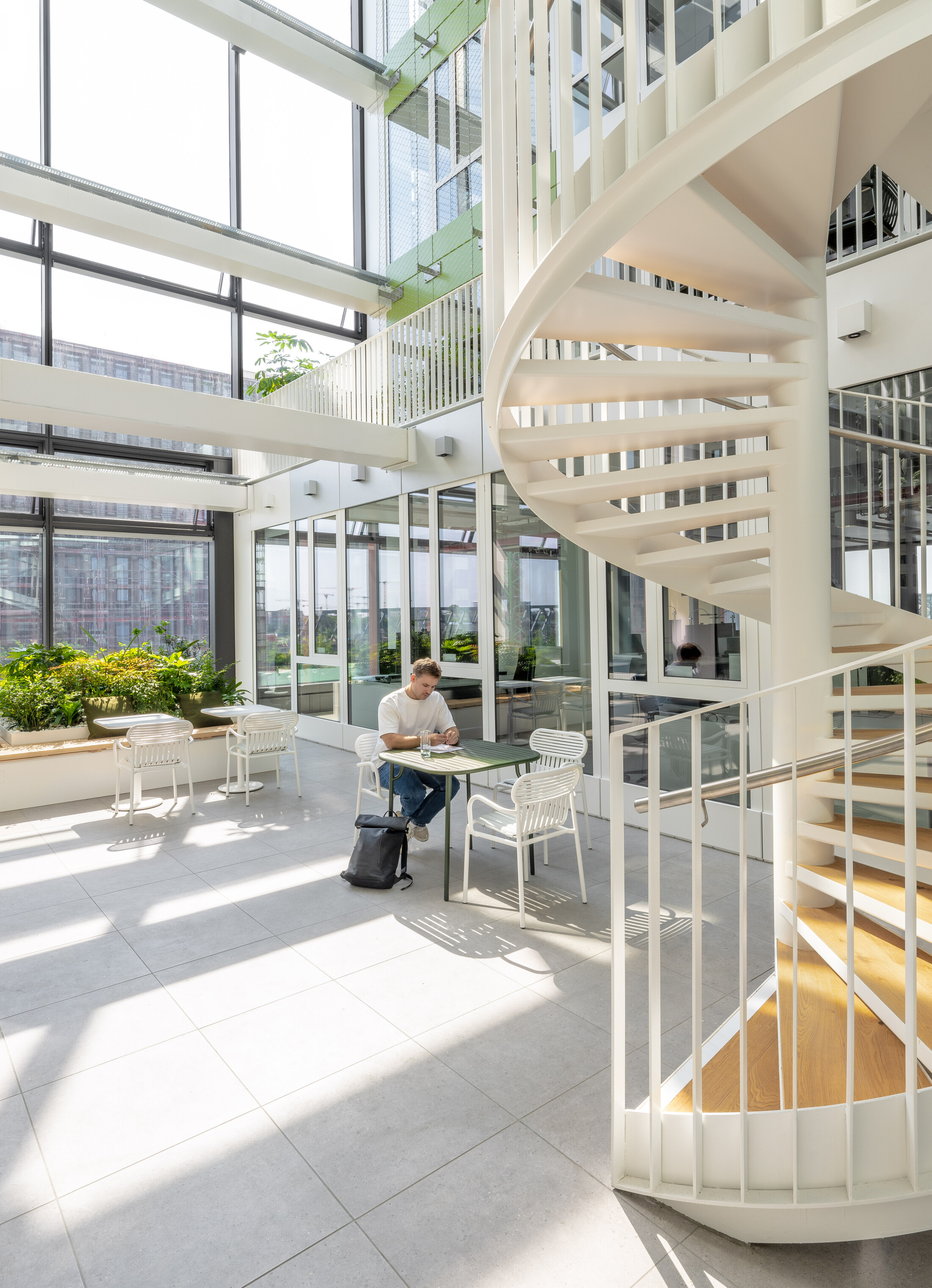 Project by Behnisch Architekturbuero, EDGE ElbSide Hamburg. Bright atrium with glass walls, a spiral staircase, and a person seated at a table working, surrounded by chairs and indoor plants.