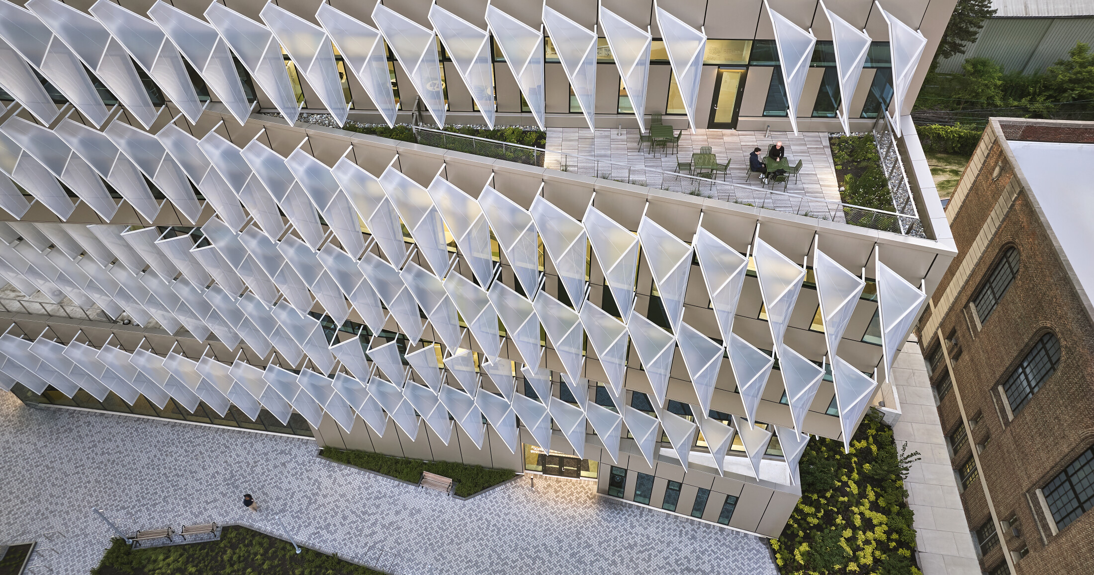 Project by Behnisch Architekturbuero, Penn VLEST. Aerial view of modern building with glass facade and angled fins, featuring a rooftop terrace with seating and people, next to a brick building.