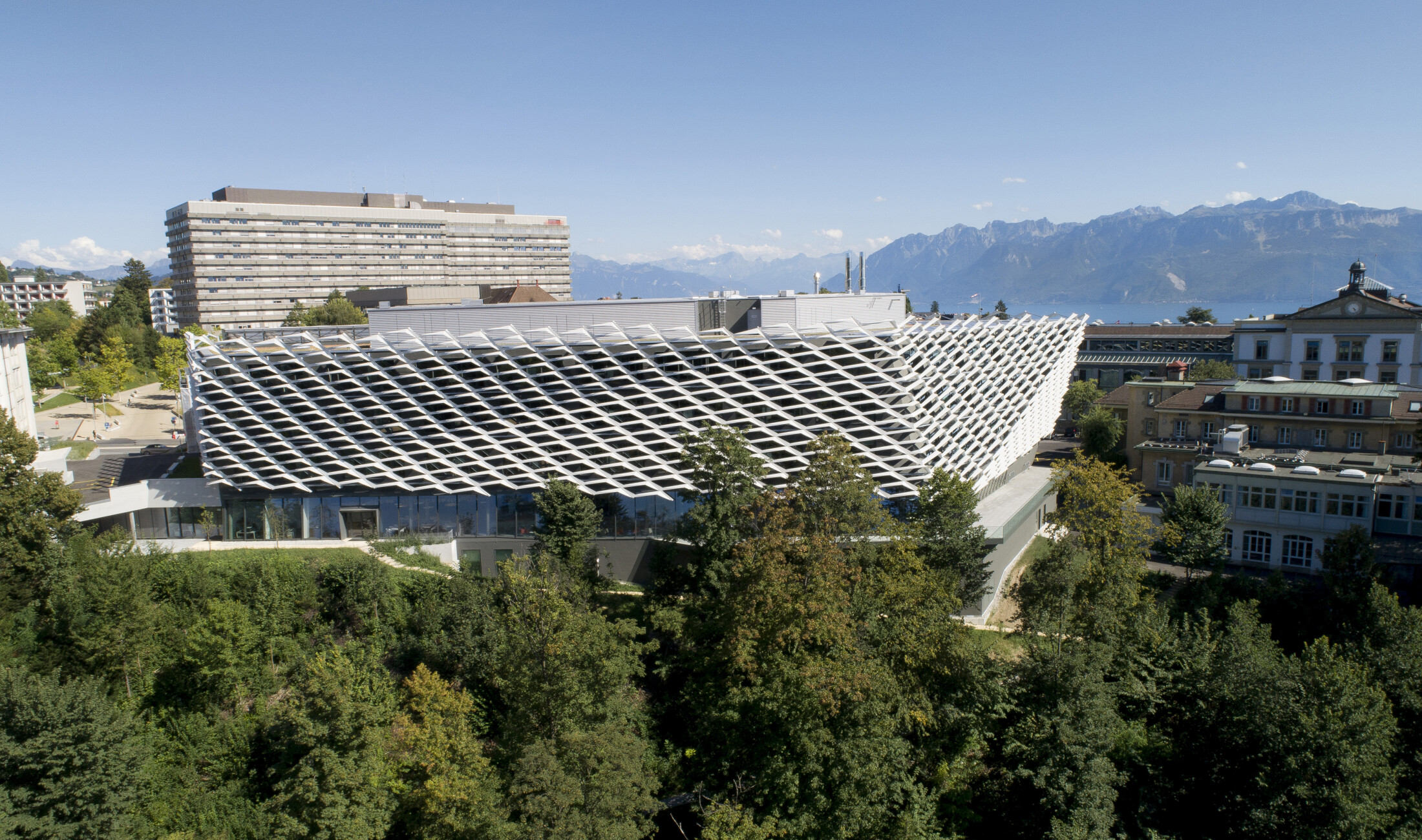 Project by Behnisch Architekturbuero, AGORA Pôle de recherche sur le cancer. Aerial view of a lakeside city with dense buildings. The large modern structure in the foreground, and mountains across the water under a clear sky.