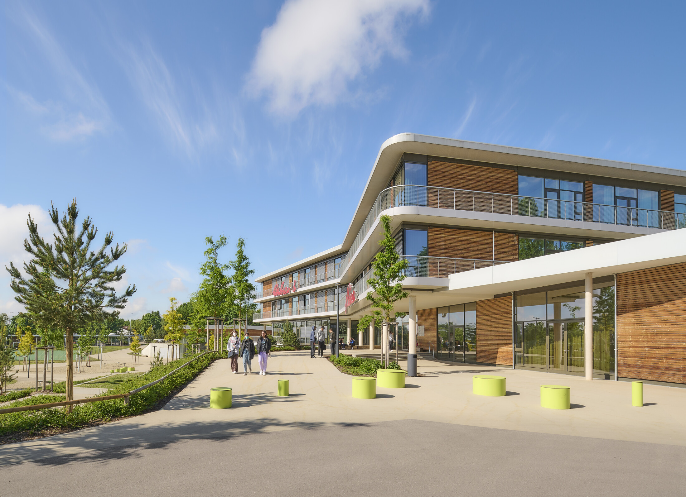 Project by Behnisch Architekturbuero, Anna-Pröll Secondary School and Sports Hall. Modern building with glass and wood facade, curved balconies, and a walkway with people and small green bollards under a blue sky.