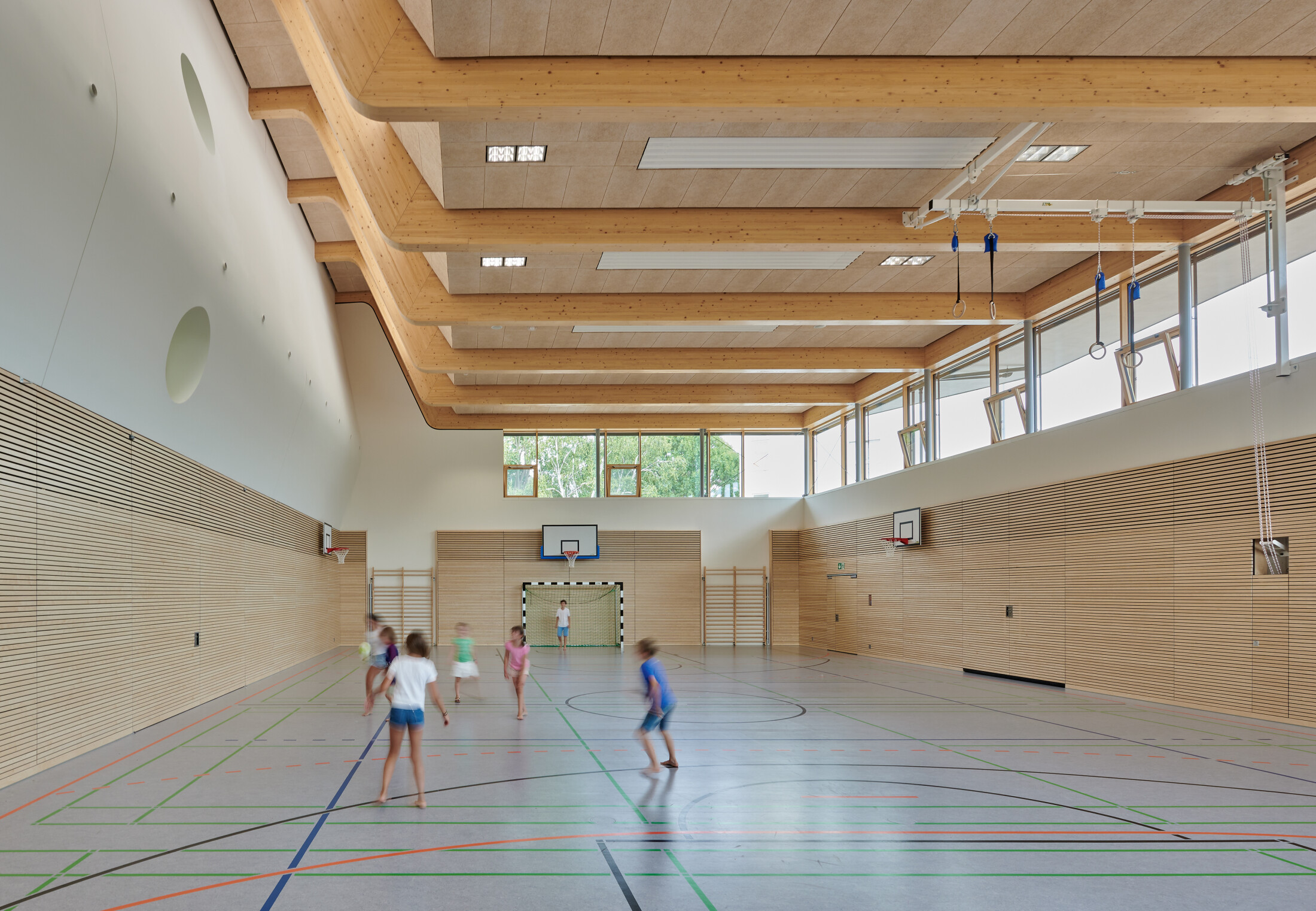 Project by Behnisch Architekturbuero, Ludwig-Weber School. School gym with wooden walls and high ceiling, children running and playing on marked court, basketball hoops and goal at far end.