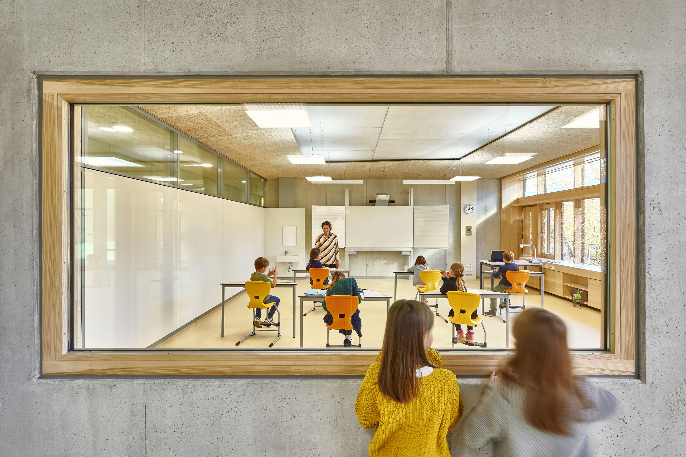 Project by Behnisch Architekturbuero, Primaryschool Infanteriestraße. View through a large window into a classroom where a teacher instructs seated children, while two students watch from the corridor outside.
