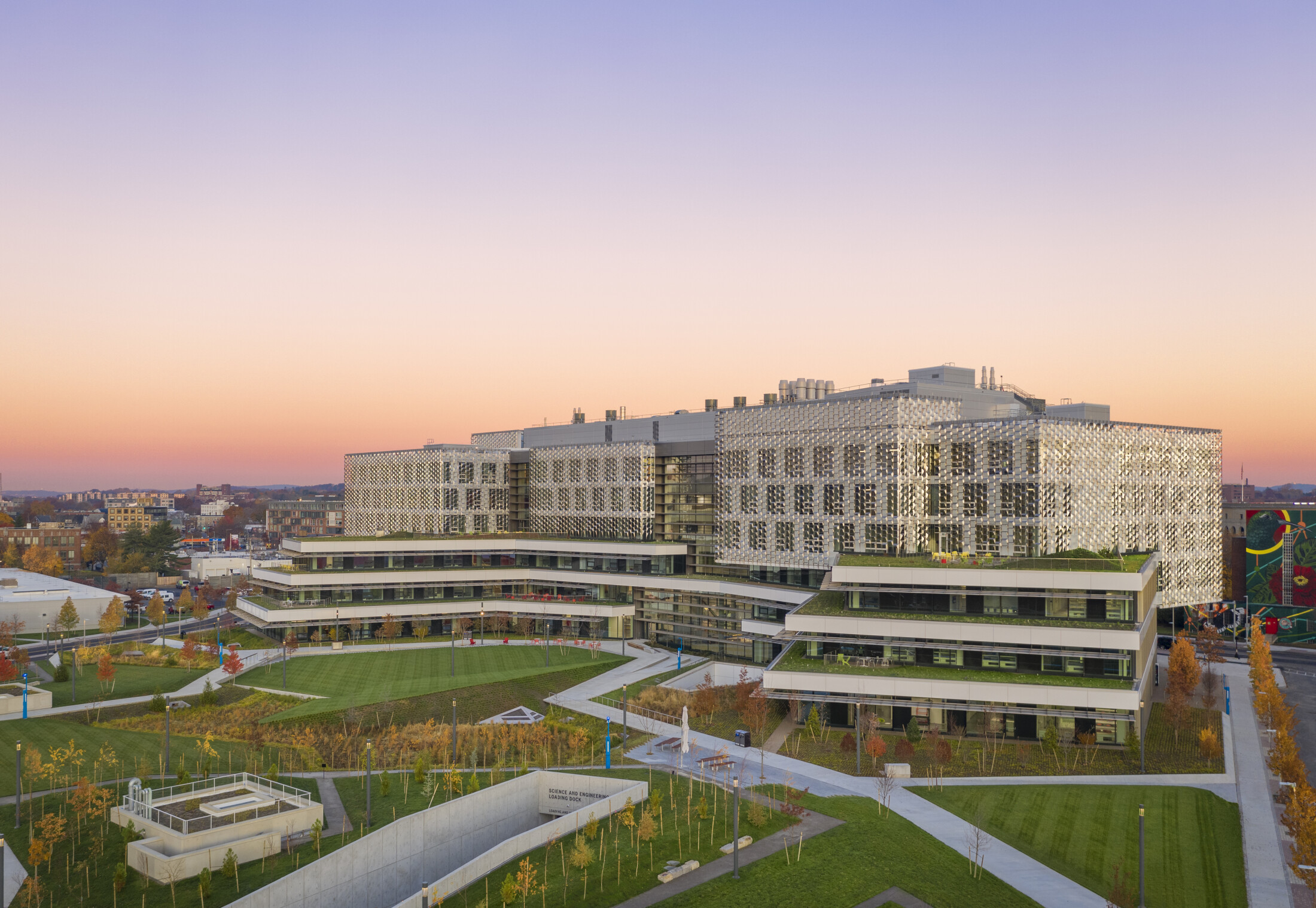 Project by Behnisch Architekturbuero, Harvard University Science and Engineering Complex (SEC). Large, modern campus building with terraced levels and patterned facade, surrounded by well-maintained lawns and paths in an evening atmosphere.